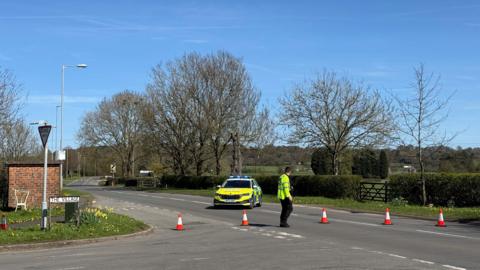 A road with orange traffic cones across it width, with a police car parked in the centre and a police officer wearing a high visibility jacket standing by the cordon. There is a brick bus shelter on the left hand side of the image and a hedgerow with trees on the right hand side.
