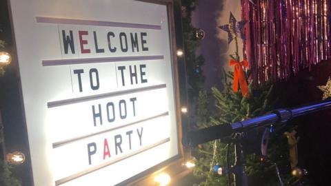 An illuminated sign which reads 'welcome to the hoot party' is surrounded by light bulbs and next to a Christmas tree with a red bow and star on top.