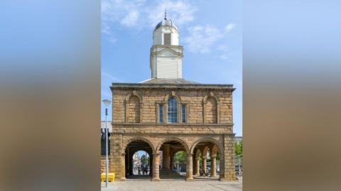 The Old Town Hall which is a stone building with an archways into the bottom floor which is open. The second floor has one central arched window and two bricked up arched windows on either side. There is a white wooden turret on the roof. The sky above is blue with a few wispy clouds. 