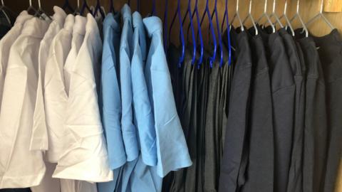White and light blue polo shirts are hung up on hangers on a rail to the left. On the right are trousers and black jumpers hung up on the same rail.