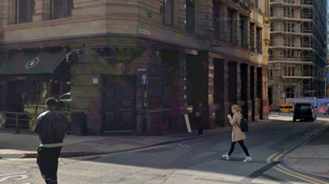 A one way road in the centre of a city. On the left there is a bar with large double doors and black awning with a logo of a bar.