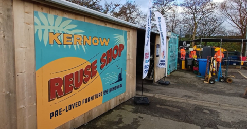 The outside of a Kernow Reuse Shop in Cornwall. It is a wooden building with a big logo and sign on the outside with two flags either side of the front entrance. There are some workers in the background wearing orange coats looking at some bins.
