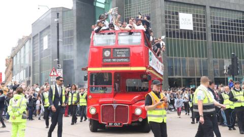 Red open top London style bus with football players on the top cheering, driving down a street lined with supporters as well as multiple men in high-viz vests