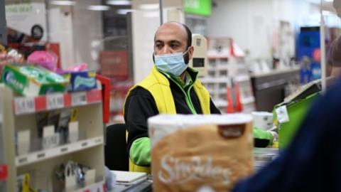 A shop worker wearing a protective face covering to combat the spread of the coronavirus, serves customers at an Asda supermarket in London