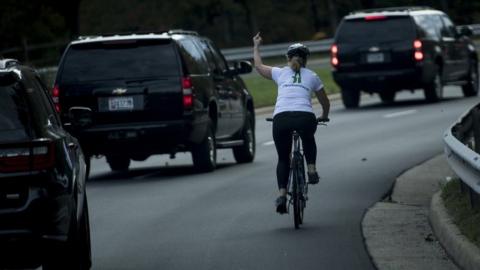 A line of motorcade cars and Juli Briskman on a cycle making a middle finger gesture