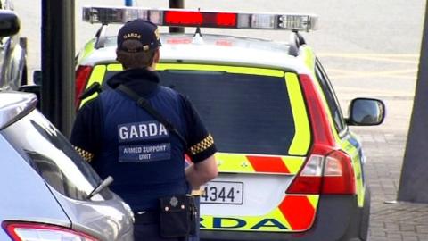 A Garda officer in a bullet proof vest stands on patrol by a police car