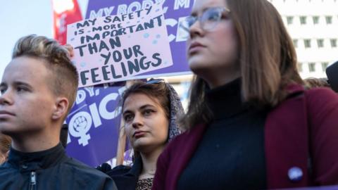 A protester holds a sign saying "my safety is more important than your feelings"