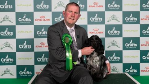 Man with big green rosette with small, black cocker spaniel sat next to him