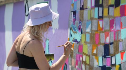 Georgie Webster wearing a lilac hat painting a wall in colourful squares