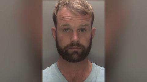 Man with brown hair and beard wearing light-coloured shirt in police mugshot