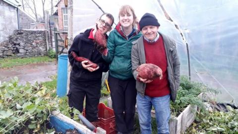 Image of two people holding sweet potatoes outside of a white polytunnel on Elm Tree Farm