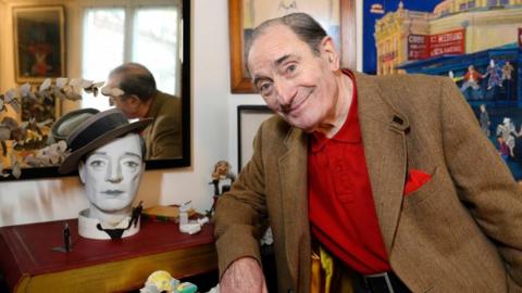 Pierre Etaix poses for a photo beside a face mask in his home