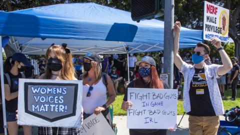 People stand on a street corner waving placards that read "Women's voices matter" and "fight bad guys in game, fight bad guys in real life"
