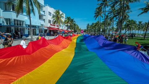 A huge multi-colored flag flies over Ocean Drive as people participate in a Pride Parade in Florida