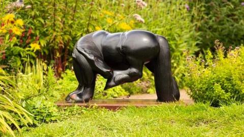 Image of the Black Mare sculpture. It is a mahogany horse, turning to nip at a fly on its rear. The sculpture has been photographed amongst grass and flowers.