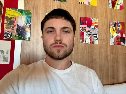 Christian Lotz has a dark brown beard and hair and is wearing a white T-shirt. He is looking directly at the camera. There are six brightly coloured red, and green portrait shaped collage-style artworks on the wall behind him, which has a brown timber-effect.