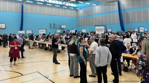 A picture of the count for Durham County Council.  Rows of tables have been laid out in a sports hall where people are seated and counting the votes. The room is filled with people awaiting the result.