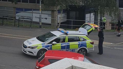 A female police officer closes the boot of a police car that is parked in the middle of a road to block traffic, with two teenage boys walking on the pavement behind her.