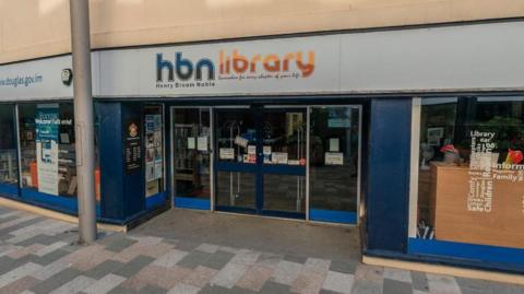 The entrance to the library featuring a sign with blue and orange writing on a white background that says HBN Library.  You can see books through the glass doors and posters are also on display.