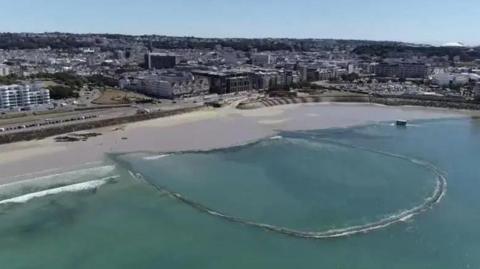 A u-shaped wall encloses sea water off a beach in St Aubin's Bay with a built up St Helier town centre in the background.