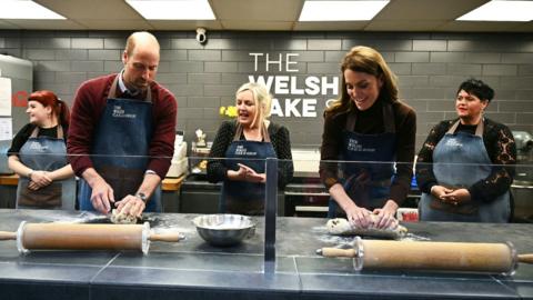 The Prince and Princess of Wales wearing aprons next to each other in a Welsh cake shop while kneading dough