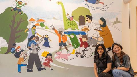 Two women sitting in front of a colourful mural installed at a children's hospice which features parents, children, a dog and a dinosaur walking around a park. 