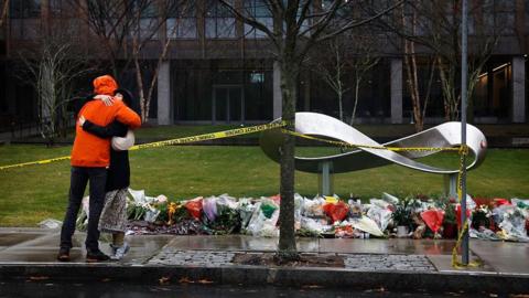 An image showing a memorial for the victims of the Brown University shooting outside a campus building where the attack took place. Dozens of flowers are placed in front of an infinity sign scultpure. In front of the memorial are a thin pole and a tree, with caution tape wrapped around them. On the left of the frame are two people embracing in front of the memorial. 