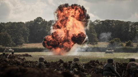 A plume of smoke and fire on a field in the background with people acting as soldiers on the floor in trenches in the foreground