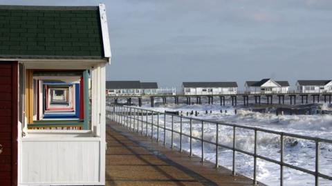 A series of colourful beach huts on the promenade in Southwold, with the huts on the left of the picture, the sea and waves crashing on the right and a pier in the distance.