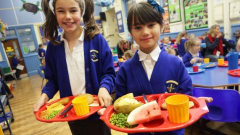 Two girls stand in a dining hall holding red trays with yellow cups on. They are wearing school uniforms and smiling at the camera. On their trays are a mixture of food including peas, beans, chips, jacket potato and watermelon. 