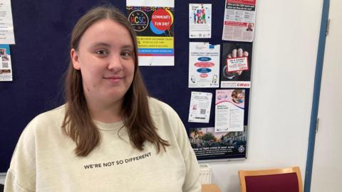 Holly Sampson with long brown hair and a "we're not so different" top in front of a noticeboard