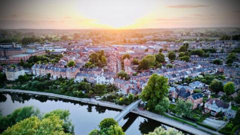 An aerial view of Shrewsbury, in the bottom of the frame is a river with a footbridge suspended above it. Next to the river is a long stretch of road lined with trees and other greenery. Behind that are several streets of houses and other buildings, mostly made of red brick