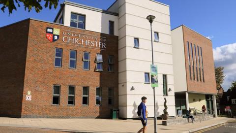 A student with a rucksack walk in front of a red brick and white paneled building with The University of Chichester written on it. 