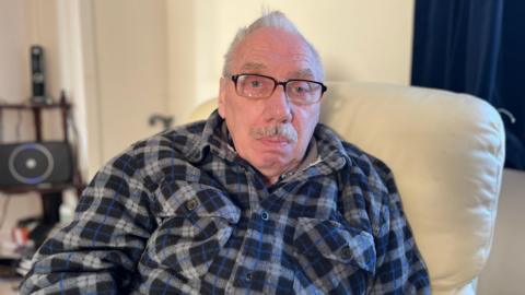 An old man sits on a cream leather armchair, looking sad. He has short grey hair and is wearing glasses and a blue and grey plaid fleece shirt jacket.