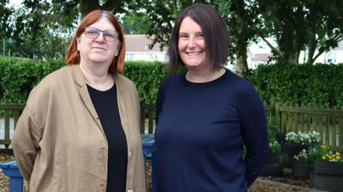 Clare Sealy and Joanne Siddall stand side by side. The pair are standing outdoors. A picket fence, flower pots a green hedge and trees are in the background.