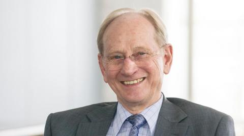 Sir David McMurtry pictured smiling at the camera. He has short, light-coloured hair and wears round frameless glasses. He also wears a light blue shirt and a darker blue tie with a diamond pattern on it, and a grey suit jacket. 