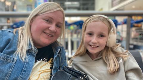 Janine and her daughter sat in the departure lounge of the airport. They both have blonde hair. Janine is wearing a blue denim jacket, and Abbie is wearing a cream hoodie and cream headphones. They are both smiling and looking at the camera