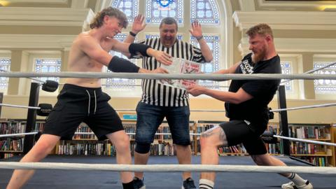 A boy in black shorts and no shirt stands on the left, a man wearing black shorts and a black T-shirt is on the right. They are both holding onto a book, fighting over it. Behind them stands a ref. All three are stood in a boxing ring. Behind them is a stained glass window and bookshelves.