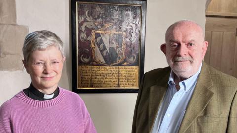 Two people standing beside a framed plaque with a decorative coat of arms, mounted on a stone wall.