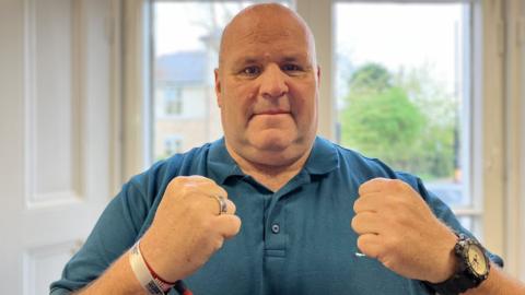 'Big John' - John Fisher stands looking at the camera with his fists clenched. He is wearing a blue polo shirt. 