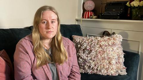 A woman in a pink shirt with long blonde hair sits on a dark green sofa. There are cushions and a blanket next to her. Behind her you can see a typewriter and some roses on a shelf.