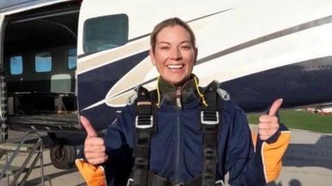 A woman wearing flight gear with her thumbs up. She is smiling at the camera. Behind her is a plane parked on the tarmac.