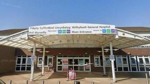 An entrance to a hospital, with a sign at the top and a car parked out front