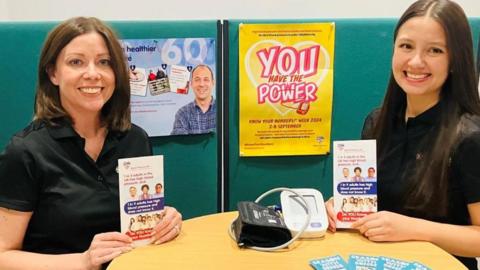 Two smiling women holding up information leaflets with blood pressure testing equipment placed on the table between them