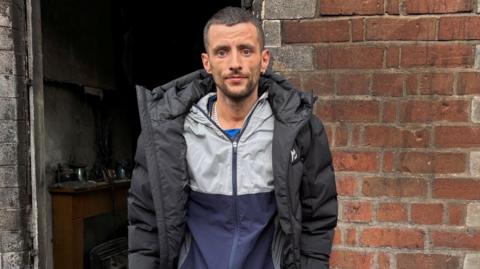 Jamie Corry stands outside of his burnt out home in east Belfast. He is looking at the camera with a neutral expression on his face. He is wearing a black puffed jacket.