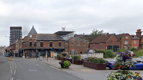 a town centre with a main road running between a row of shops and a car park