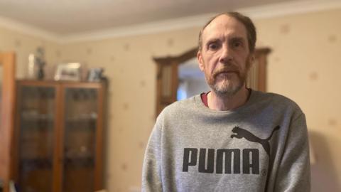 Gary Punter is wearing a grey puma sweatshirt and is standing in the back room of his rental property. He is looking at the camera and has beard. Behind him can be seen a corner, glass-fronted cupboard.