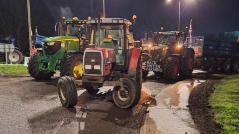 Multiple tractors are parked on a road, completely blocking access. One has a sign that says "Back British farming" on the front.