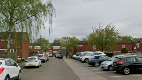 A street view image of Vauxhall Crescent with cars parked on the right hand side of the road. Trees line the road and terraced houses sit beyond the pavement.