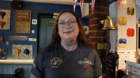 Janine Bird smiling at the camera. She had long red hair and is wearing glasses and a Guinness-branded T-shirt. She is stood inside a pub, which has colourful walls decorated with various pieces of artwork.